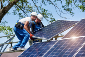 Two male roofers wearing white hard hats, installing solar panels on residential home